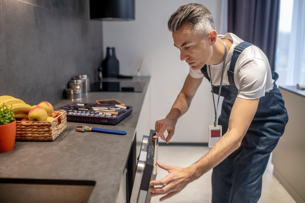 Technician in overalls repairing built-in oven in kitchen.