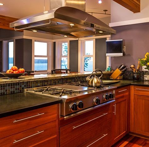 Kitchen island with gas stove, kettle, and overhead steel range hood near windows.