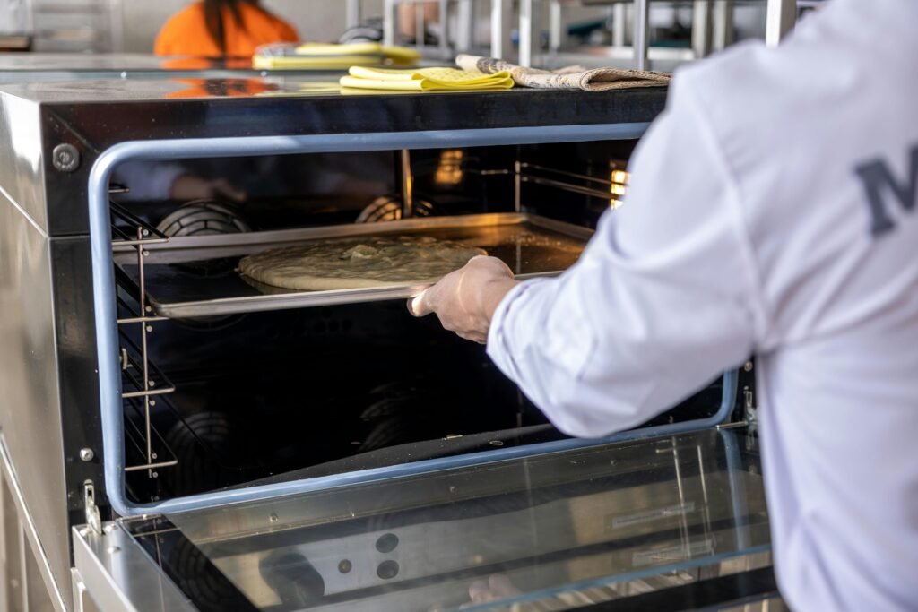 Chef in white uniform placing dough on tray inside commercial oven.