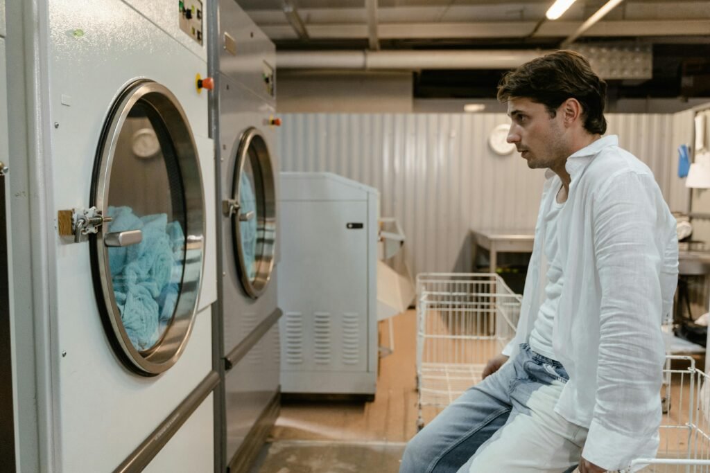 Man sitting near industrial washing machines in laundry room.
